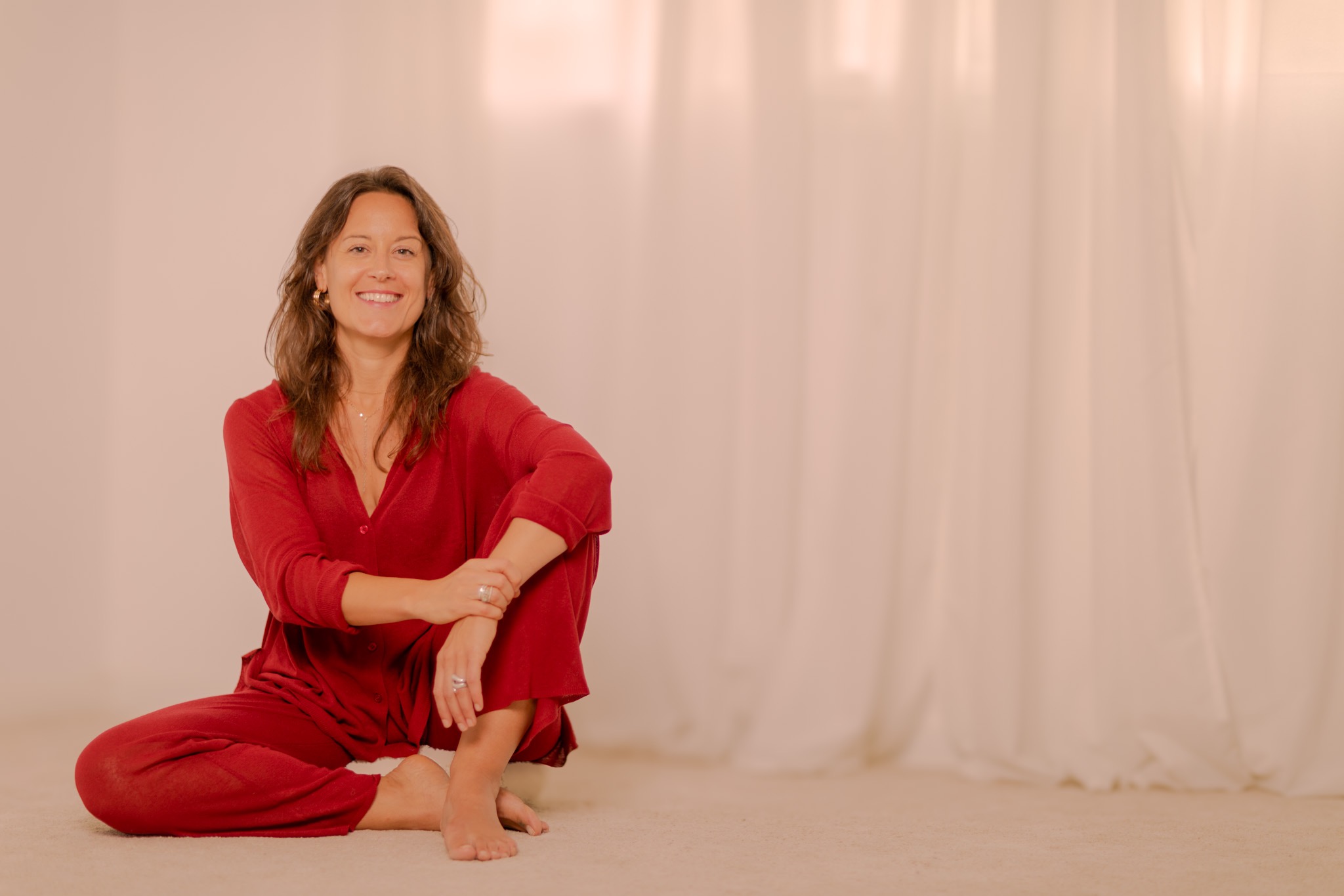 Núria Boladeres seated cross-legged on the floor in a red linen shirt, smiling warmly, soft natural light and white curtains behind her.