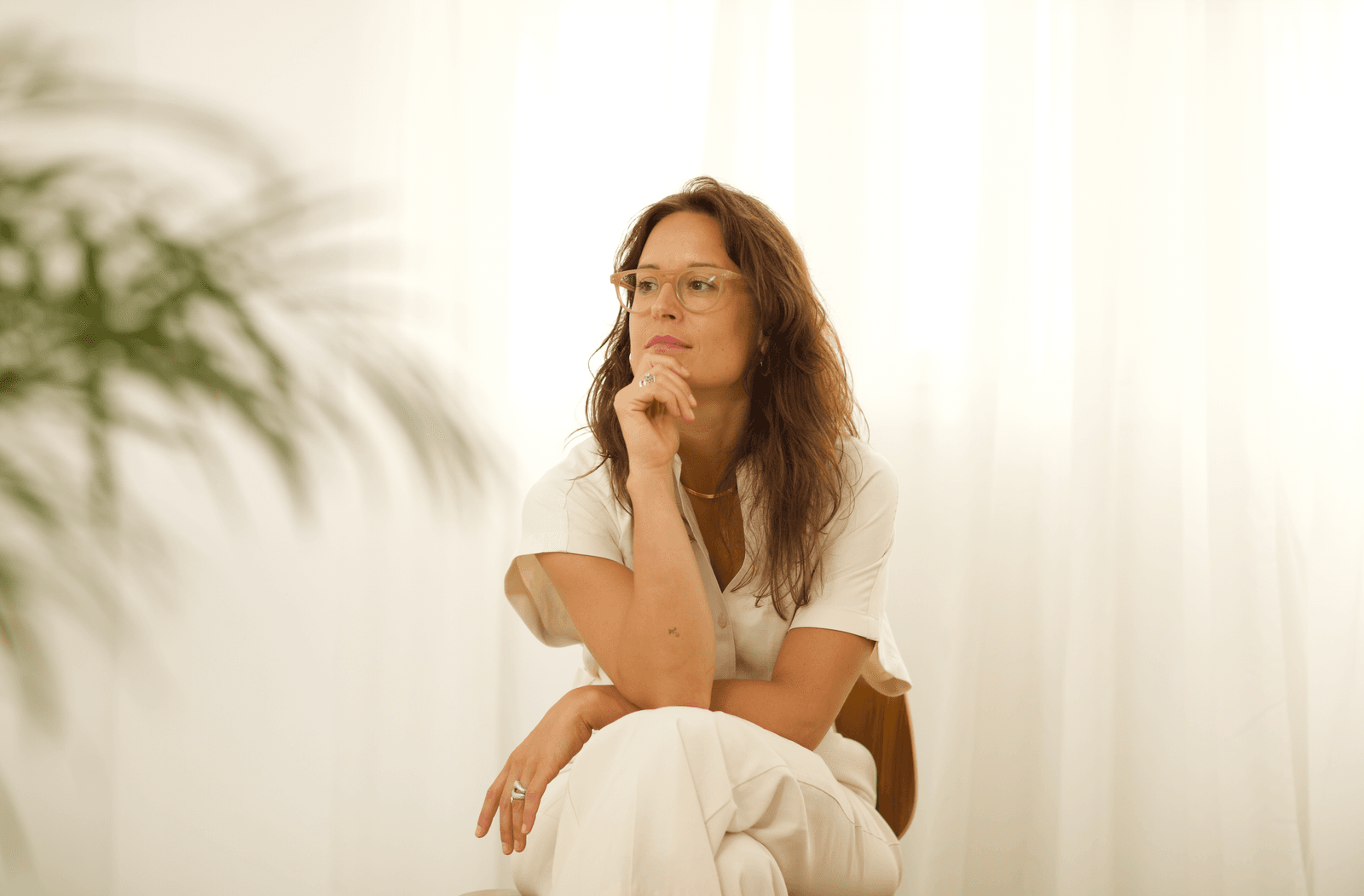 Núria seated in a bright room, hand resting at her chin, soft natural light through curtains, palm shadow falling left.