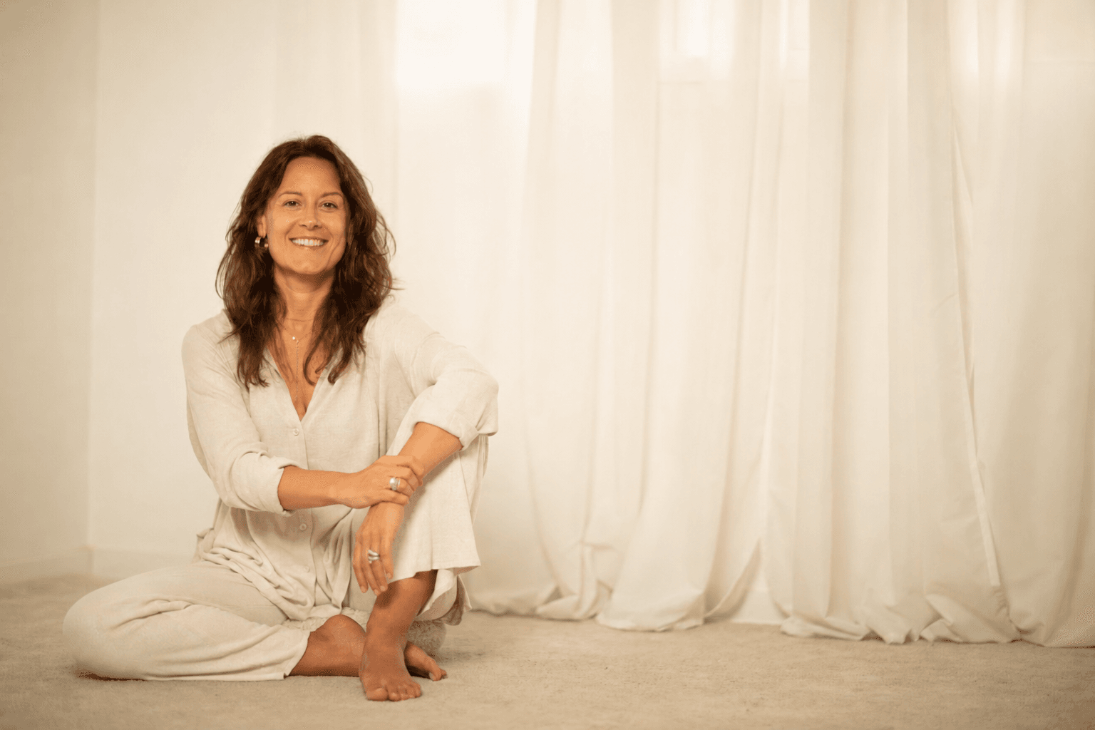 Núria Boladeres seated cross-legged on the floor in a soft cream linen shirt, smiling, bright natural light through white curtains behind her.